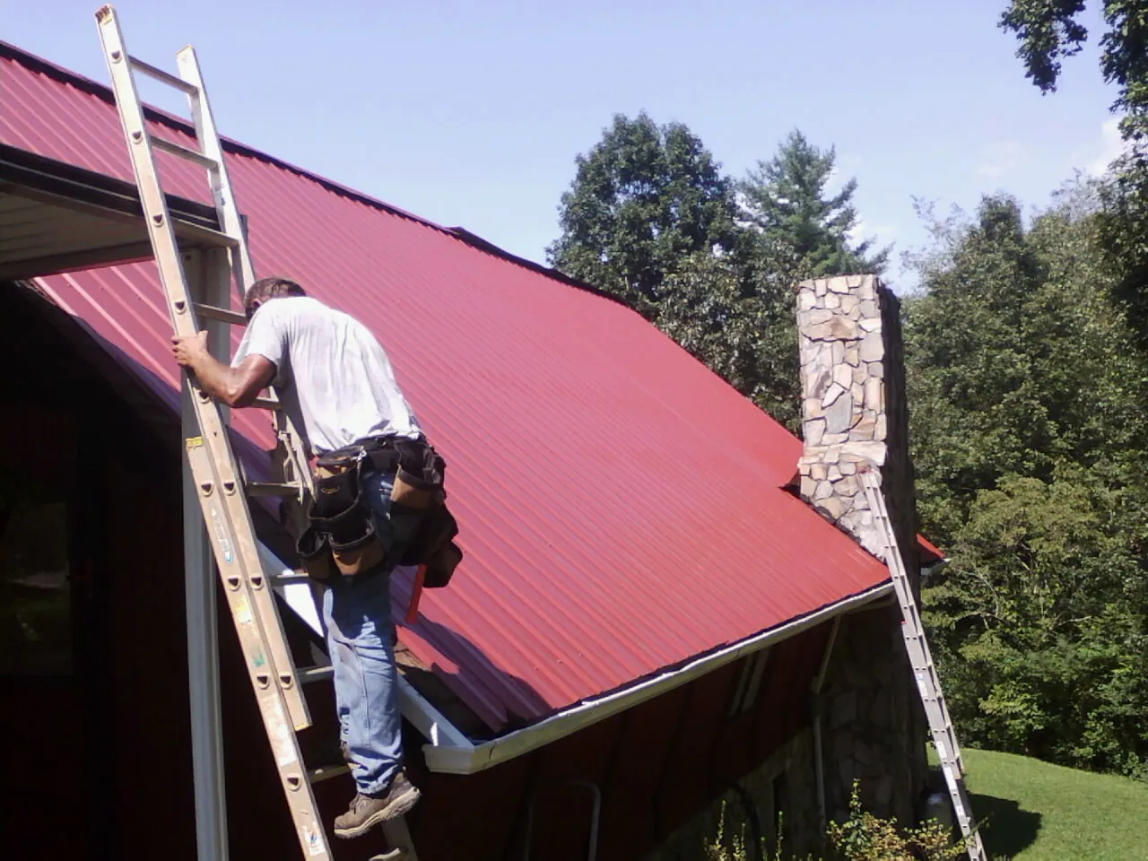 Roofer on a ladder beside a red metal roof and stone chimney — Soffit & Fascia Repair by Roofing Brothers in Campaign